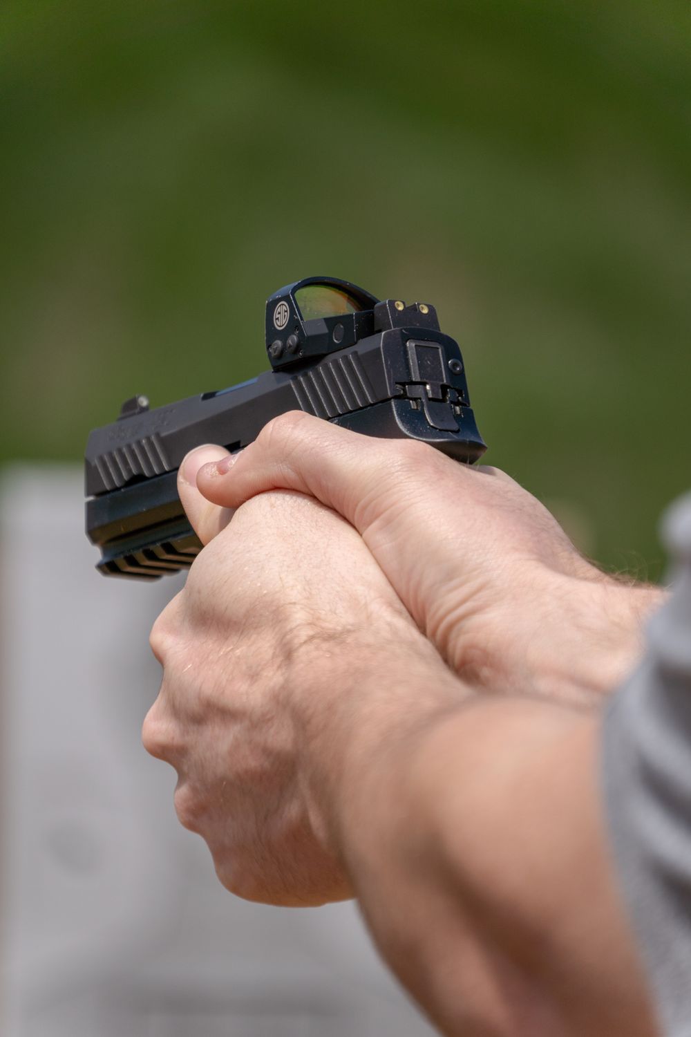 Close-up of hands gripping a black semi-automatic pistol with red dot sight outdoors.