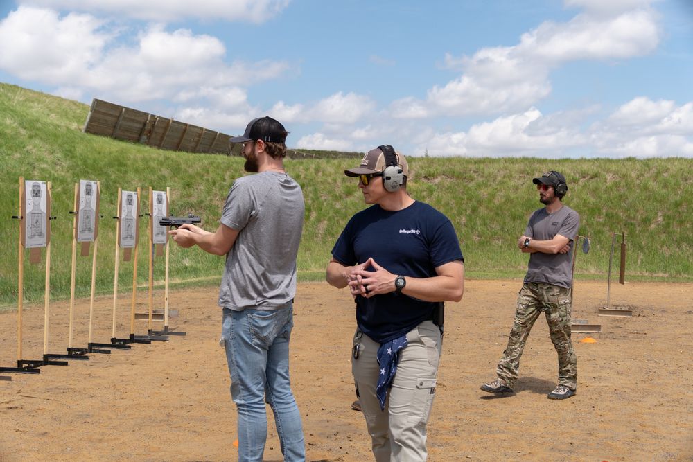 Three men at an outdoor shooting range with paper targets, wearing ear protection during a firearms training session.