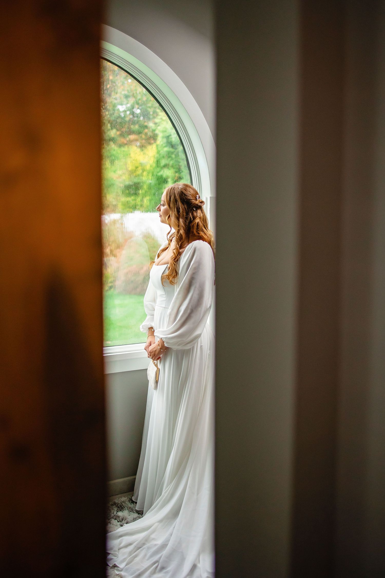A person in a flowing white wedding dress stands by an arched window, creating a serene silhouette against natural light.