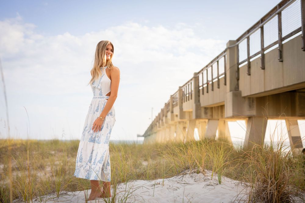 A person in a white dress stands on a sandy beach boardwalk during golden hour at sunset.
