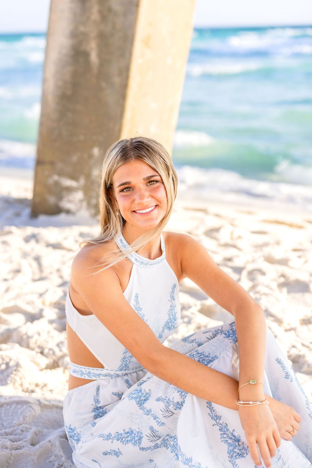 A person in a white sundress relaxes on a sandy beach near ocean waves and wooden pier posts.