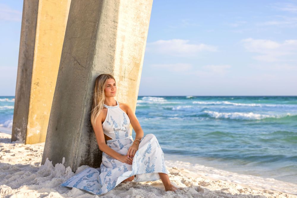 Woman in white dress sitting by stone pillars on tropical beach with turquoise ocean waves in background.