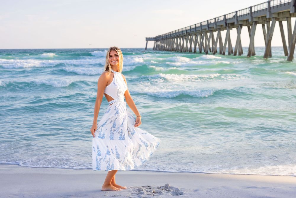 A person in a flowing white dress walks along the shoreline near a wooden pier as turquoise waves roll in.