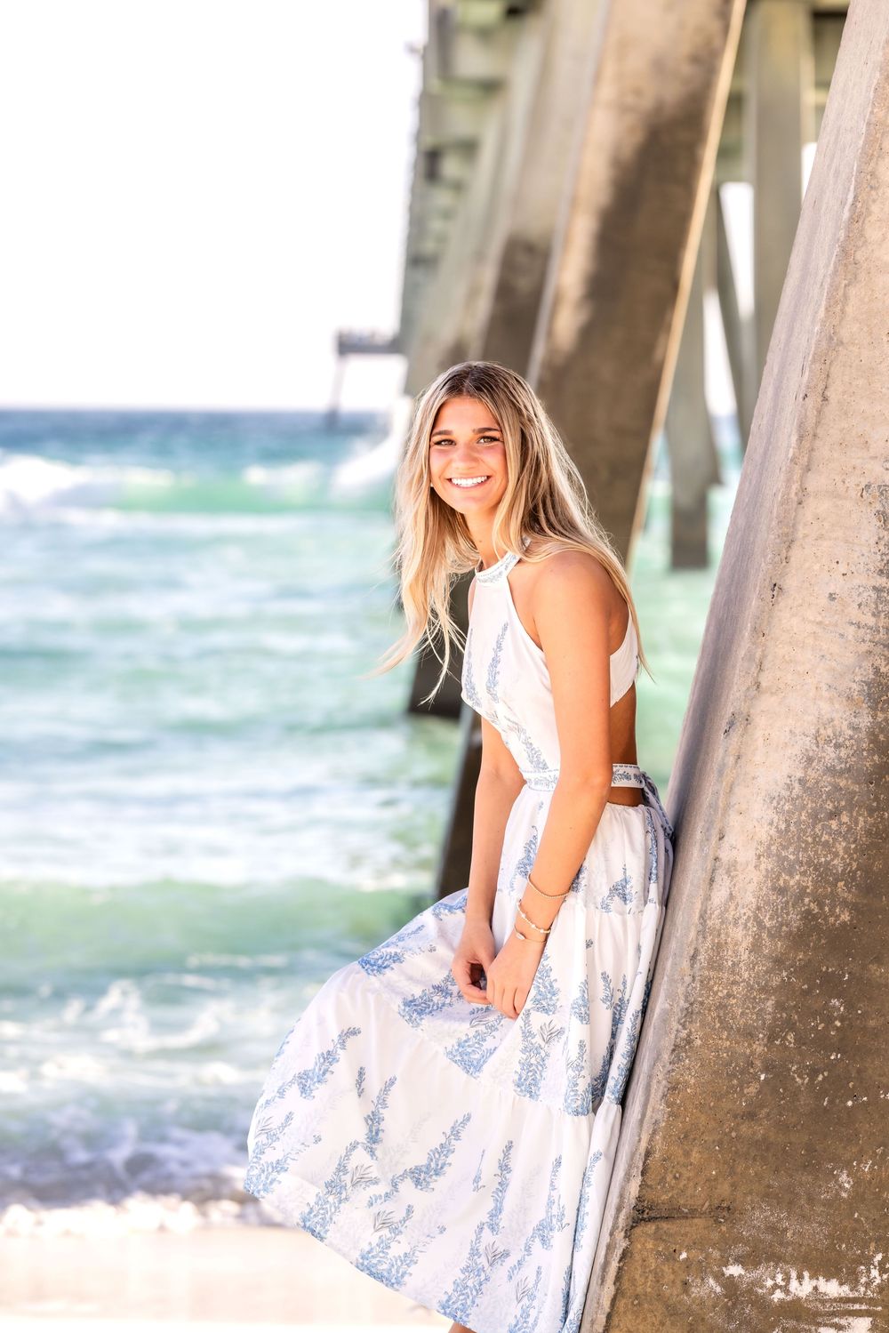 A white sundress contrasts against weathered pier posts as ocean waves crash in the background at sunset.