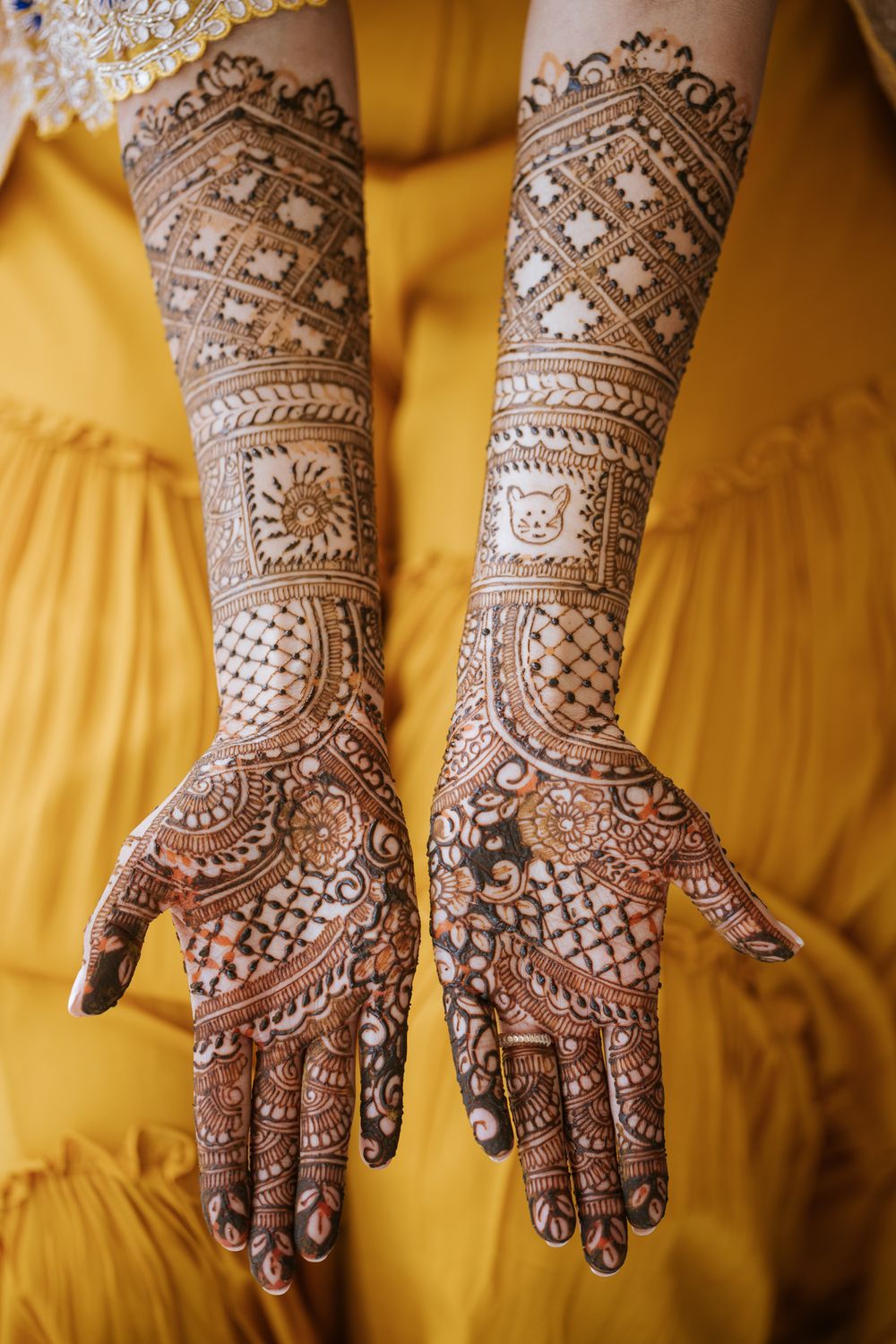 Intricate bridal mehndi design featuring detailed geometric patterns and traditional motifs on both hands against yellow fabric.