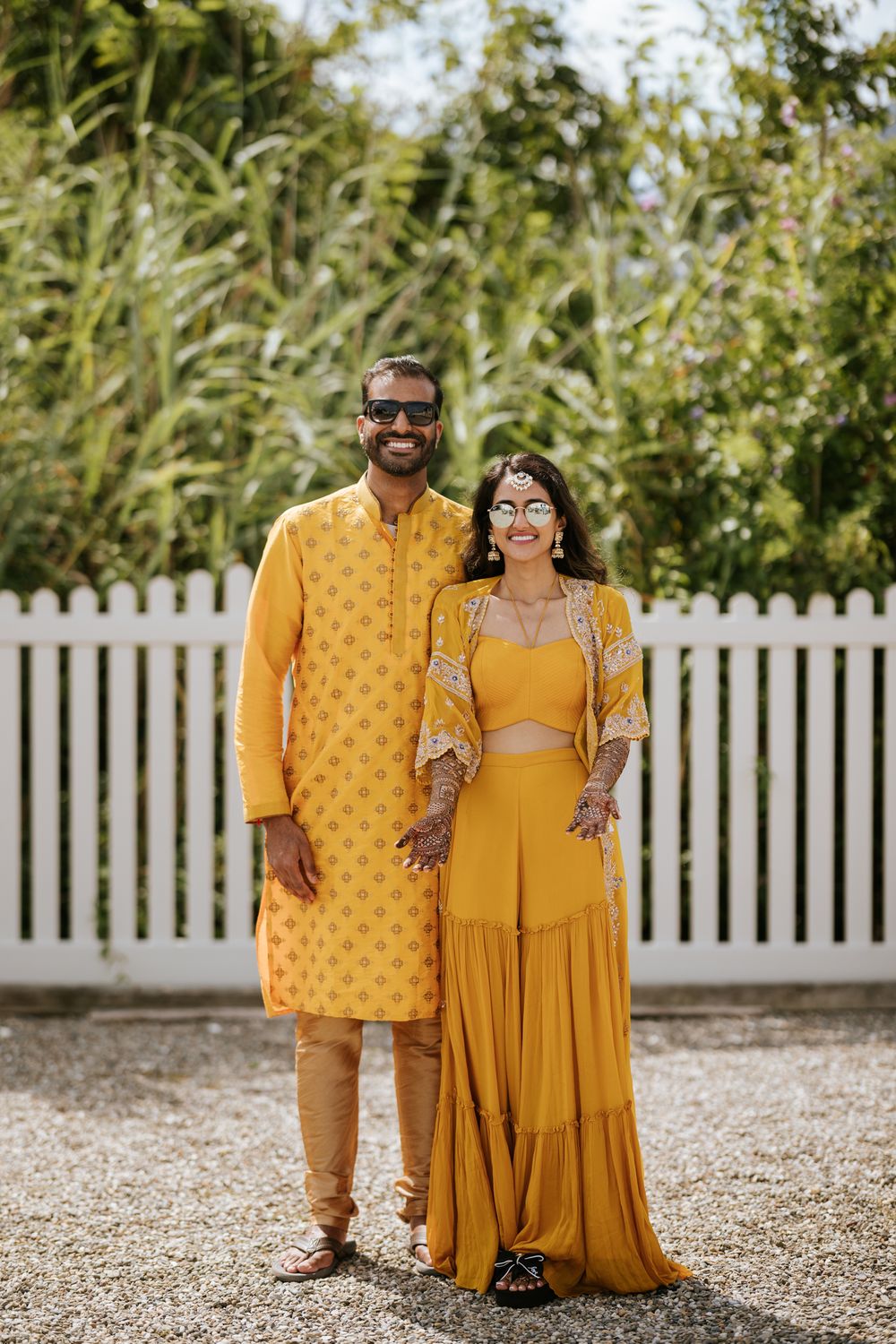 A couple in matching yellow traditional Indian outfits pose together in front of a white picket fence and greenery.