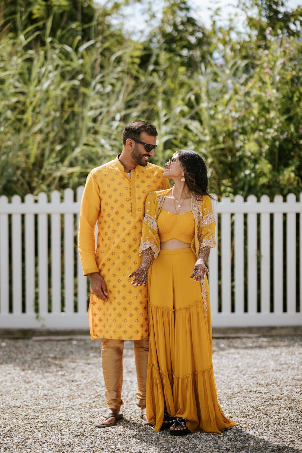 A couple in coordinating yellow Indian outfits pose together against a white picket fence and greenery backdrop.