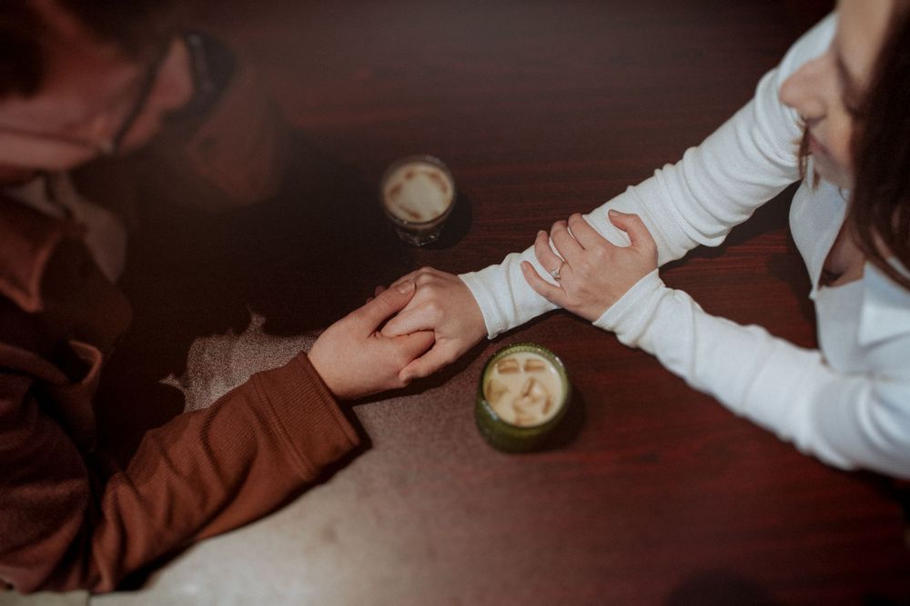 Two people hold hands across a table with coffee cups in a warm, intimate setting. Indianapolis, Indiana engagement session.
