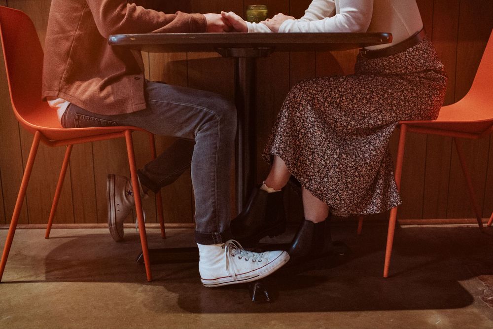 Two people sit at a small table in orange chairs, with one wearing white sneakers and the other in a floral dress. Indianapolis, Indiana