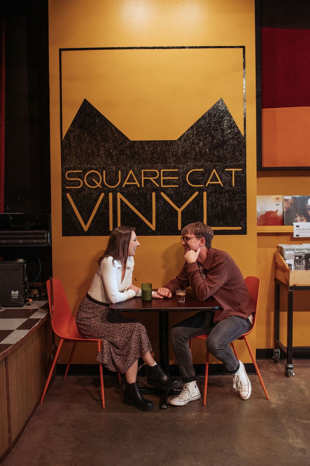 Two people sit in modern chairs having a conversation inside a cafe with yellow walls and dark artwork. Indianapolis, Indiana engagement