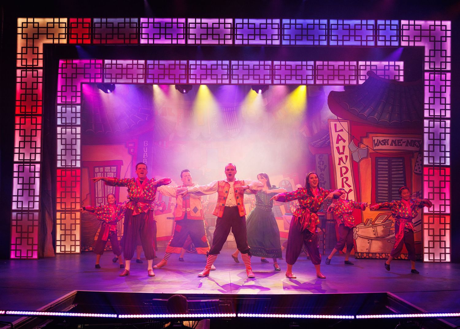 Performers dance on a colorfully lit stage with vibrant purple and red lighting during a theatrical production.