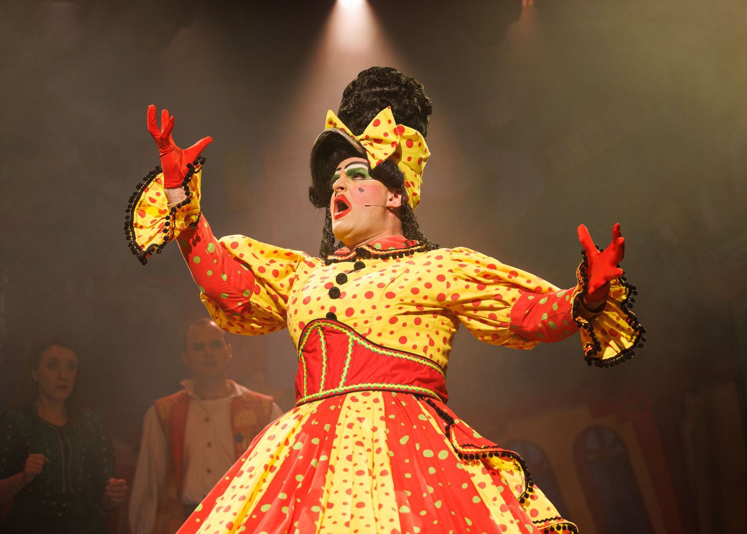 A performer in a bright yellow and red polka dot dress dances energetically on a dimly lit stage during a theatrical show.