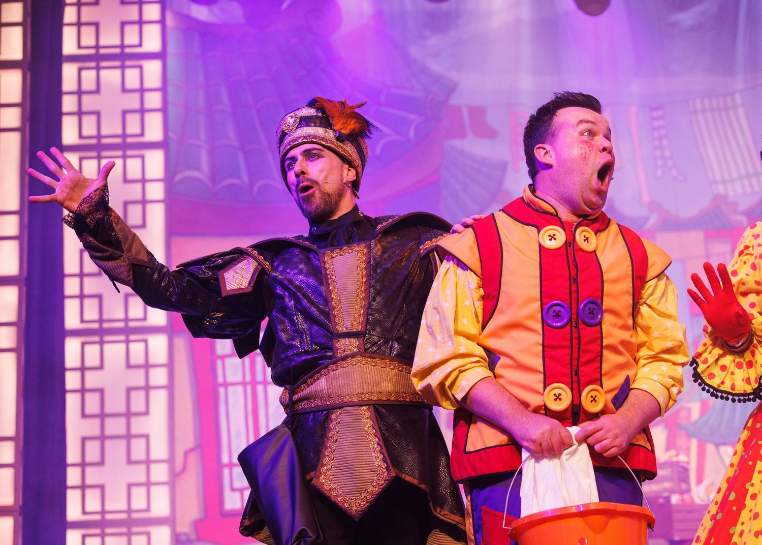 Performers in bright orange and black costumes dance energetically on a colorfully lit stage during a theatrical production.