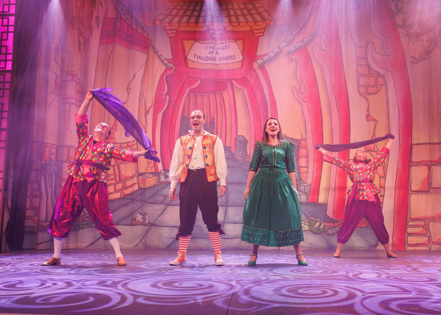 Theater cast members dancing in colorful costumes on a pink-lit stage with ornate set design.