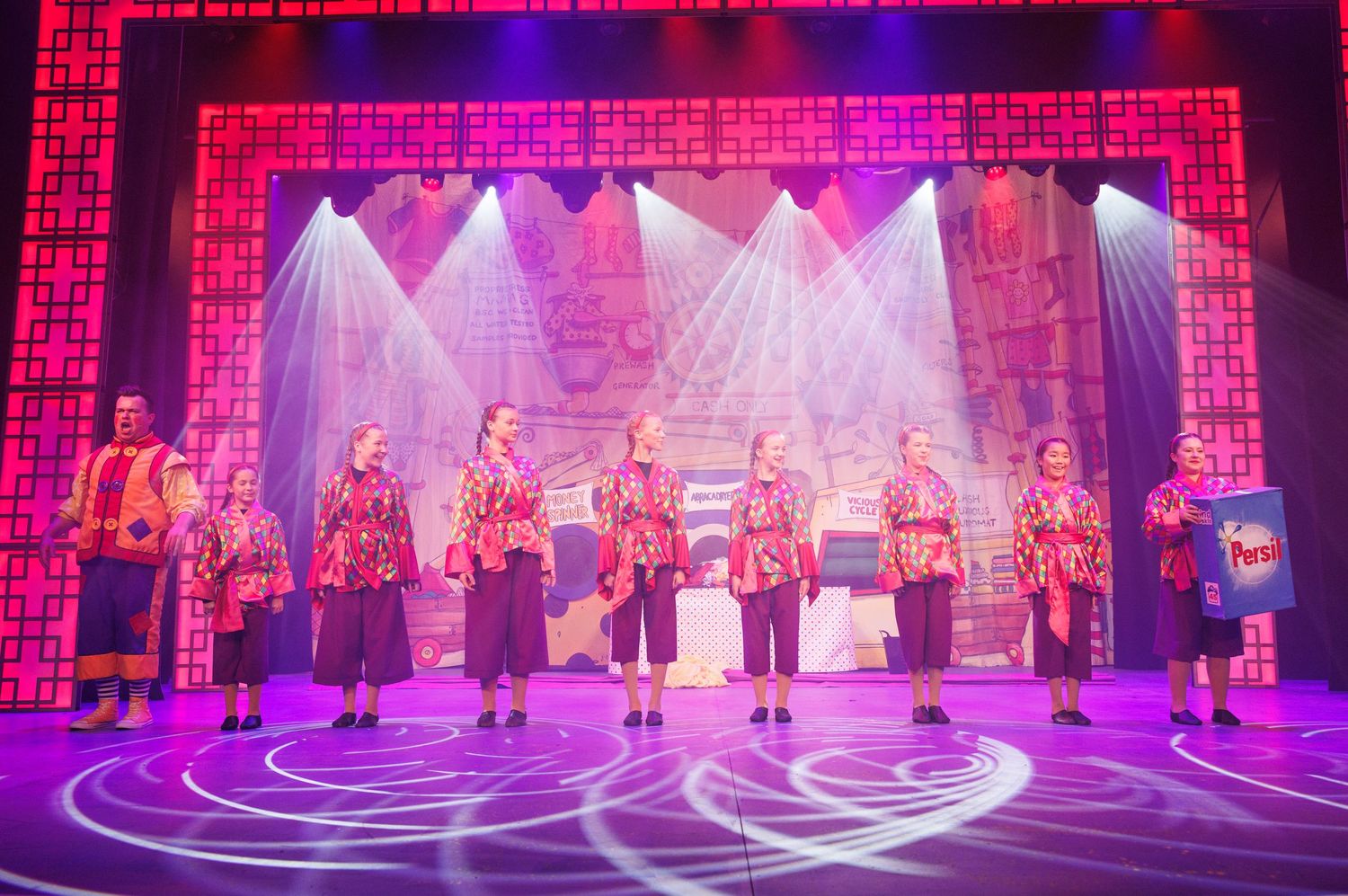 Stage performers dance in vibrant pink and purple lighting during a theatrical production.