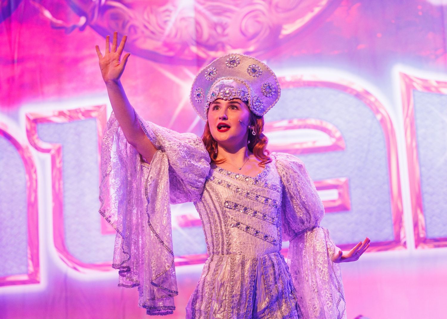 A performer in a lavender Victorian-style costume and wig gestures dramatically against a bright pink stage backdrop.