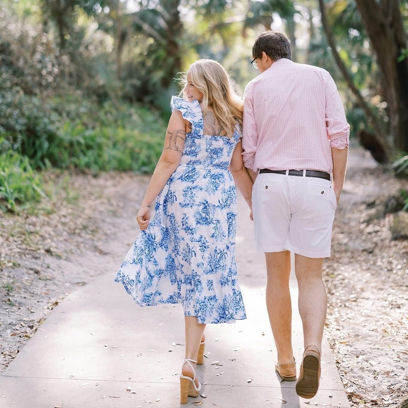 A couple walks hand in hand down a palm-lined dirt path, with her wearing a blue floral dress and him in pink and khaki.