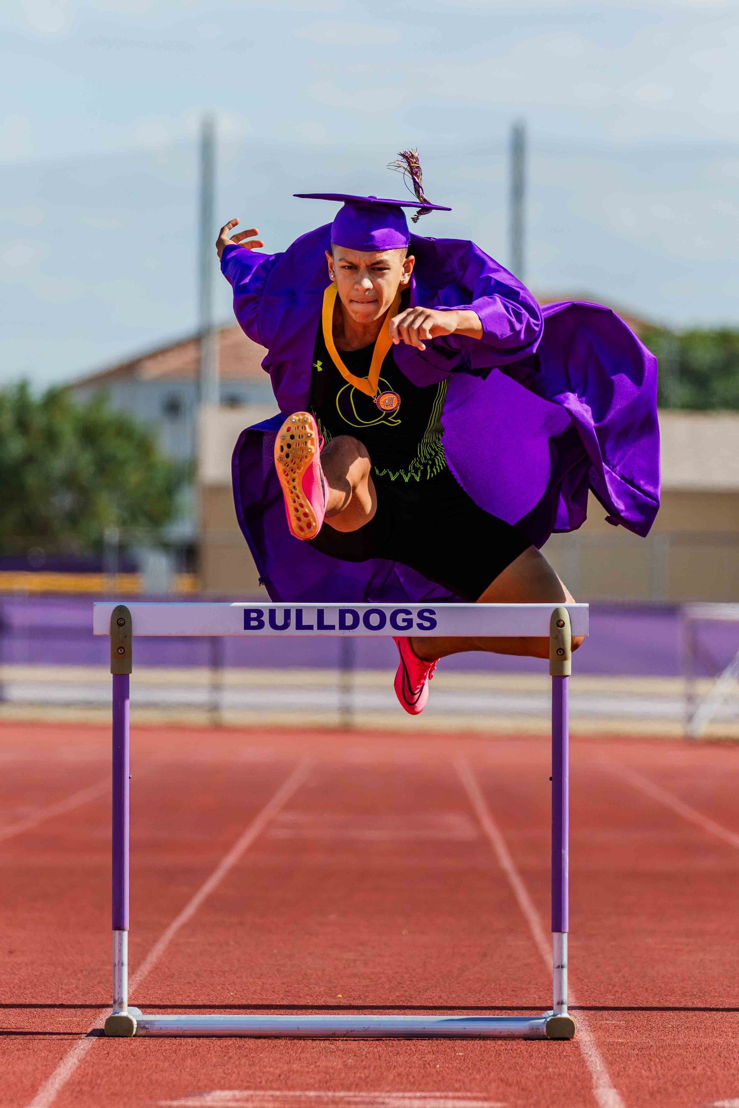 Graduate in purple cap and gown leaps over hurdle on track during celebratory photo shoot at Bulldogs stadium.