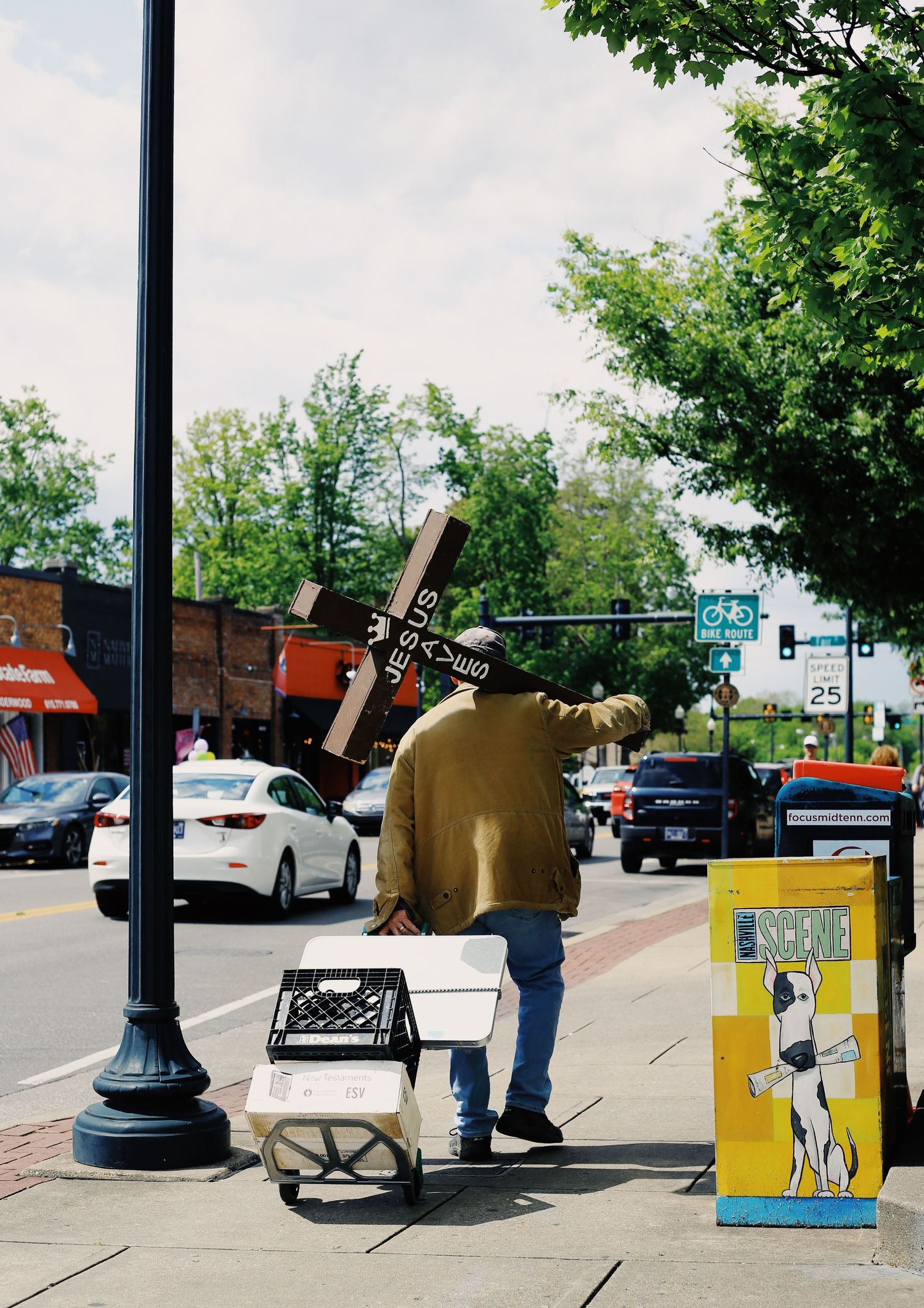 Street performer carrying a large cross and cart setup on an urban sidewalk with trees and parked cars in background.