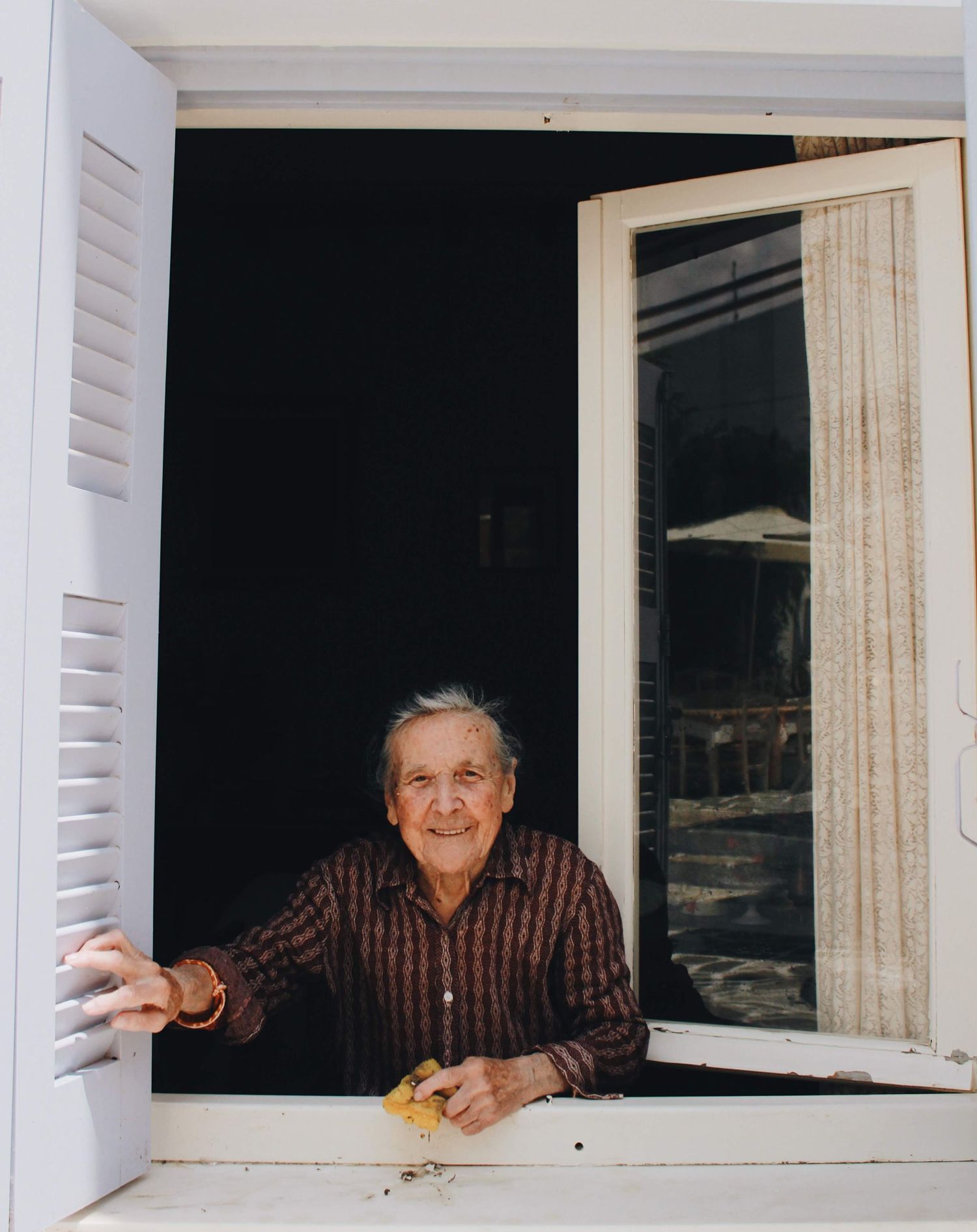Someone leans out of a white-framed window with a warm smile, wearing a plaid shirt in this nostalgic photograph.