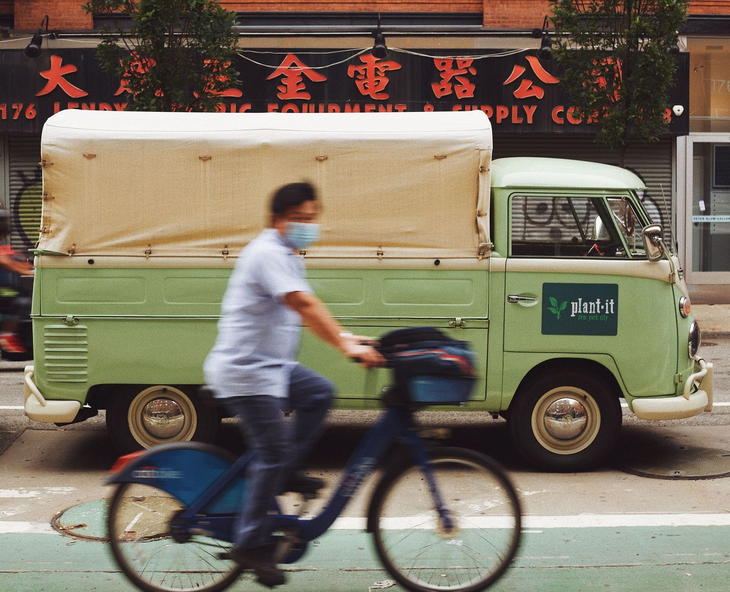 A row of vintage green delivery vans and bicycles parked along a city street in an urban setting.