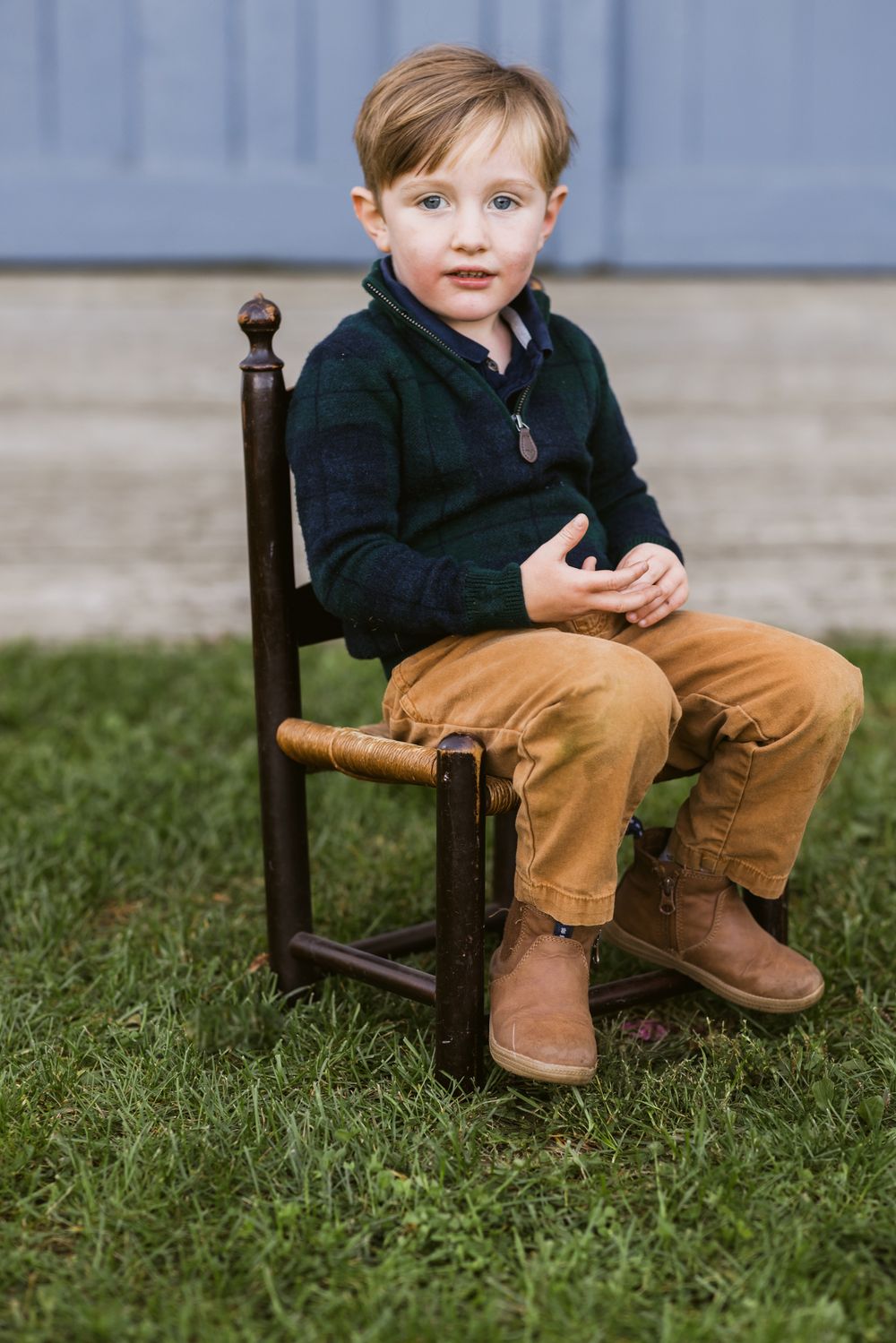 A portrait of a young boy sits on a small chair in front of pale blue door at Saltonstall Farm in Stratham, New Hampshire for family photos.