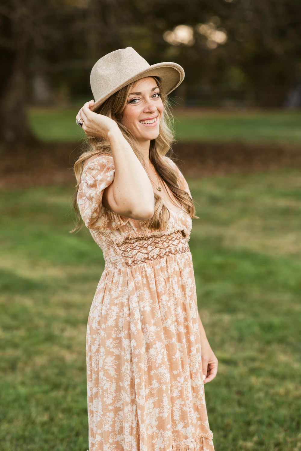 A mother wearing a tan suede hat and orange floral peasant dress poses in a lush green field for motherhood photos in the Seacoast region NH