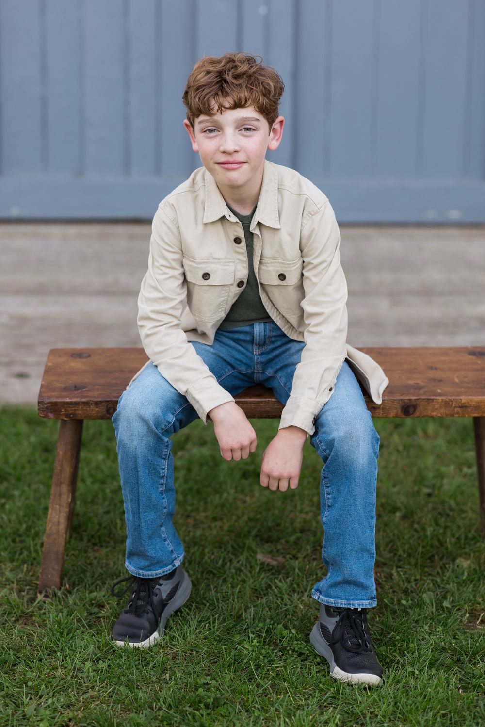 An older boy with auburn curly hair wearing cream jacket blue jeans sits on a wooden bench in front of a pastel blue door for family photos.