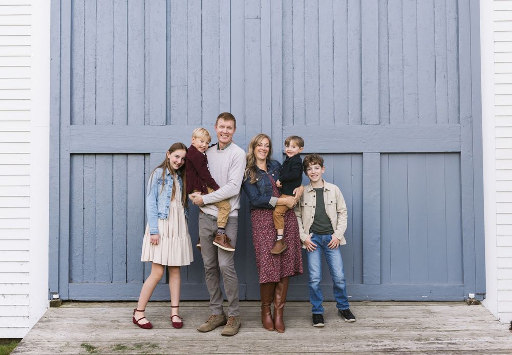 A smiling family poses in front of a pale blue door at Saltonstall Farm in Stratham, Seacoast New Hampshire for family photos.