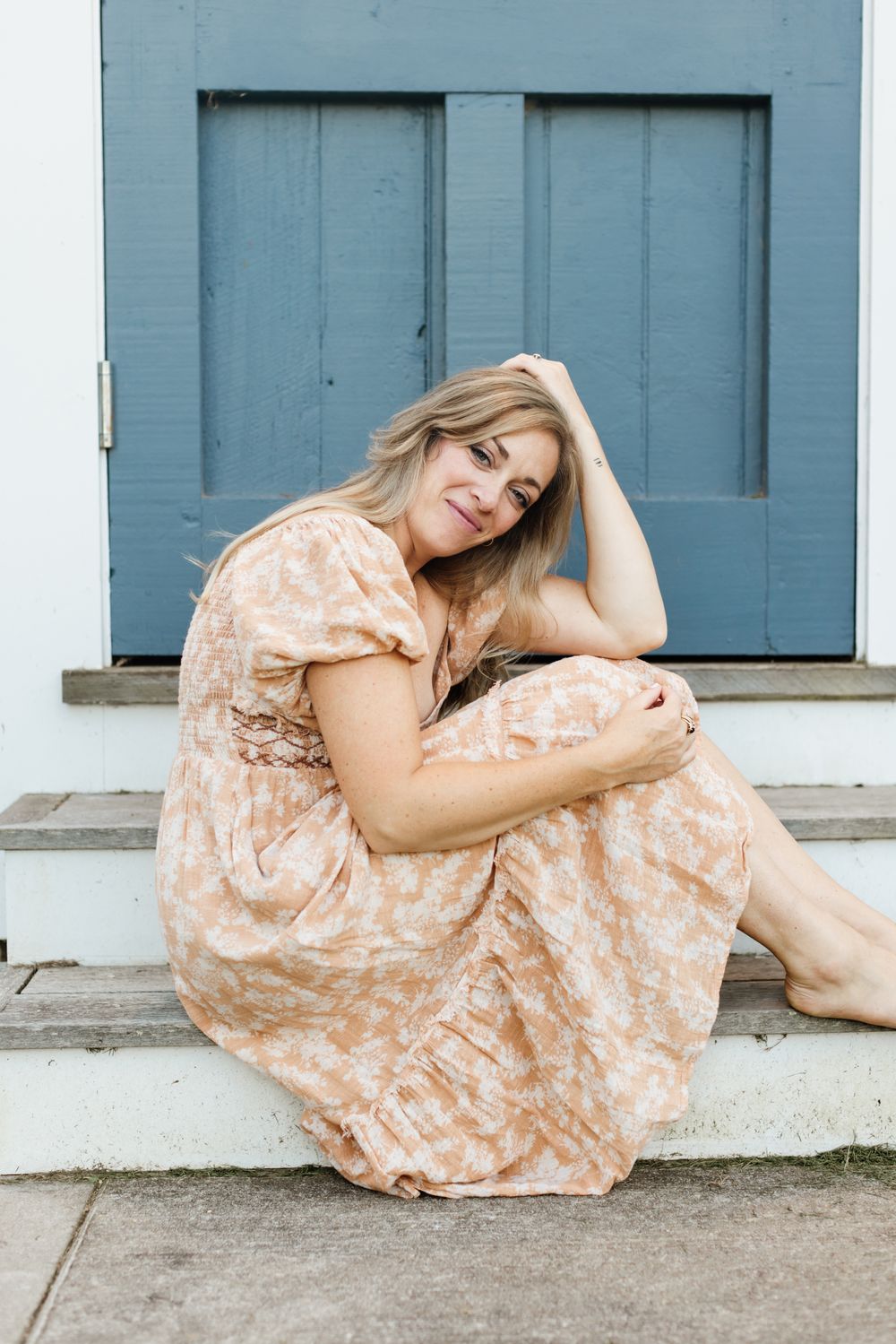 A mother wearing an orange floral dress poses and sits gracefully in front of a pastel blue door at Saltonstall Farm for a motherhood photo