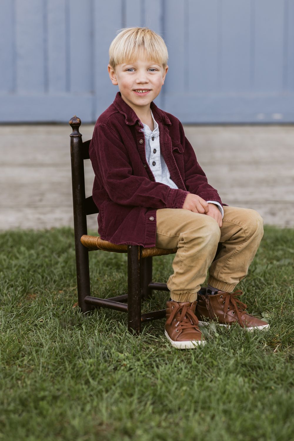 A young boy with blonde hair sits on small chair in front of a blue door at Saltonstall Farm in Stratham, New Hampshire for family photos.