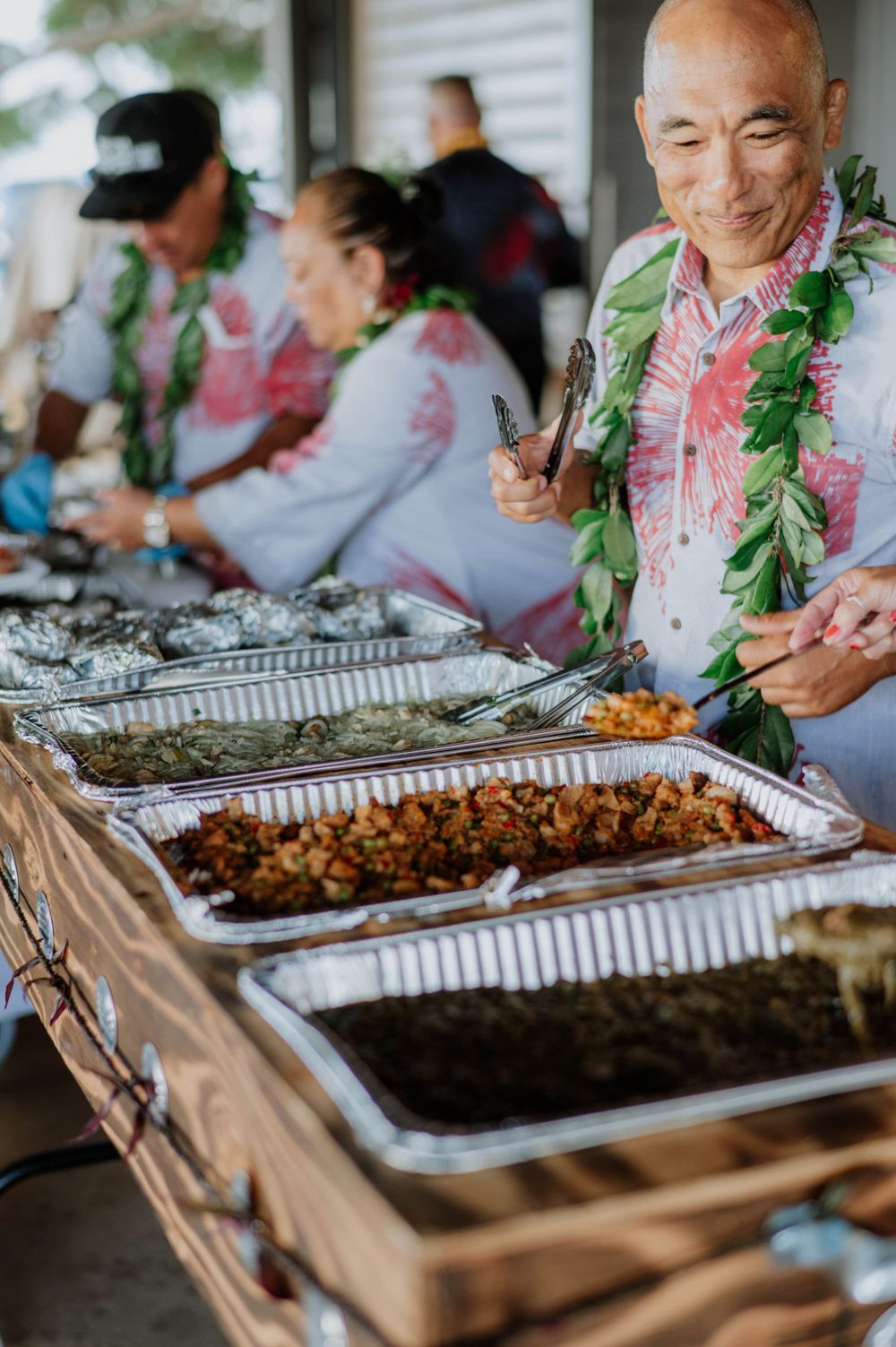 Wedding guests wearing traditional leis serve food from a buffet line.
