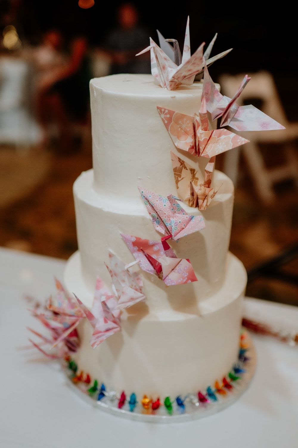 Three-tiered white wedding cake decorated with pink origami paper cranes and colorful base.