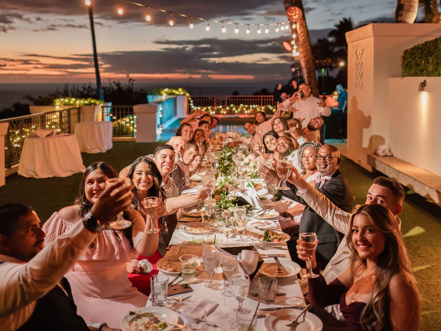 Wedding guests enjoying an outdoor reception dinner under string lights at sunset.