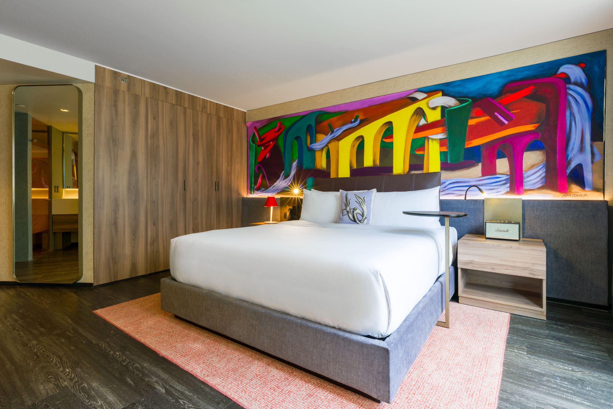 Modern hotel room interior with sleek furnishings, white bedding, and colorful abstract artwork on the wall.