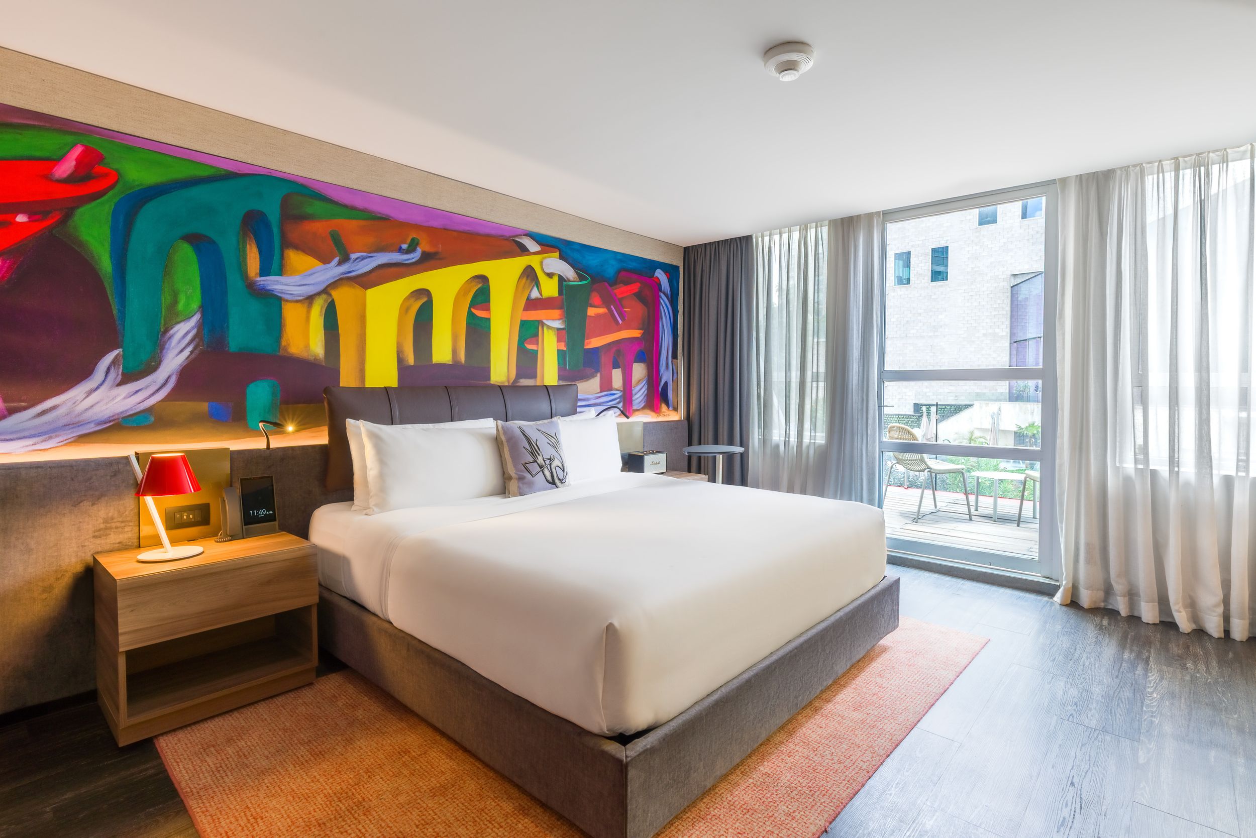 Modern hotel room interior with white bedding, large windows, and colorful abstract wall art creating a vibrant atmosphere.