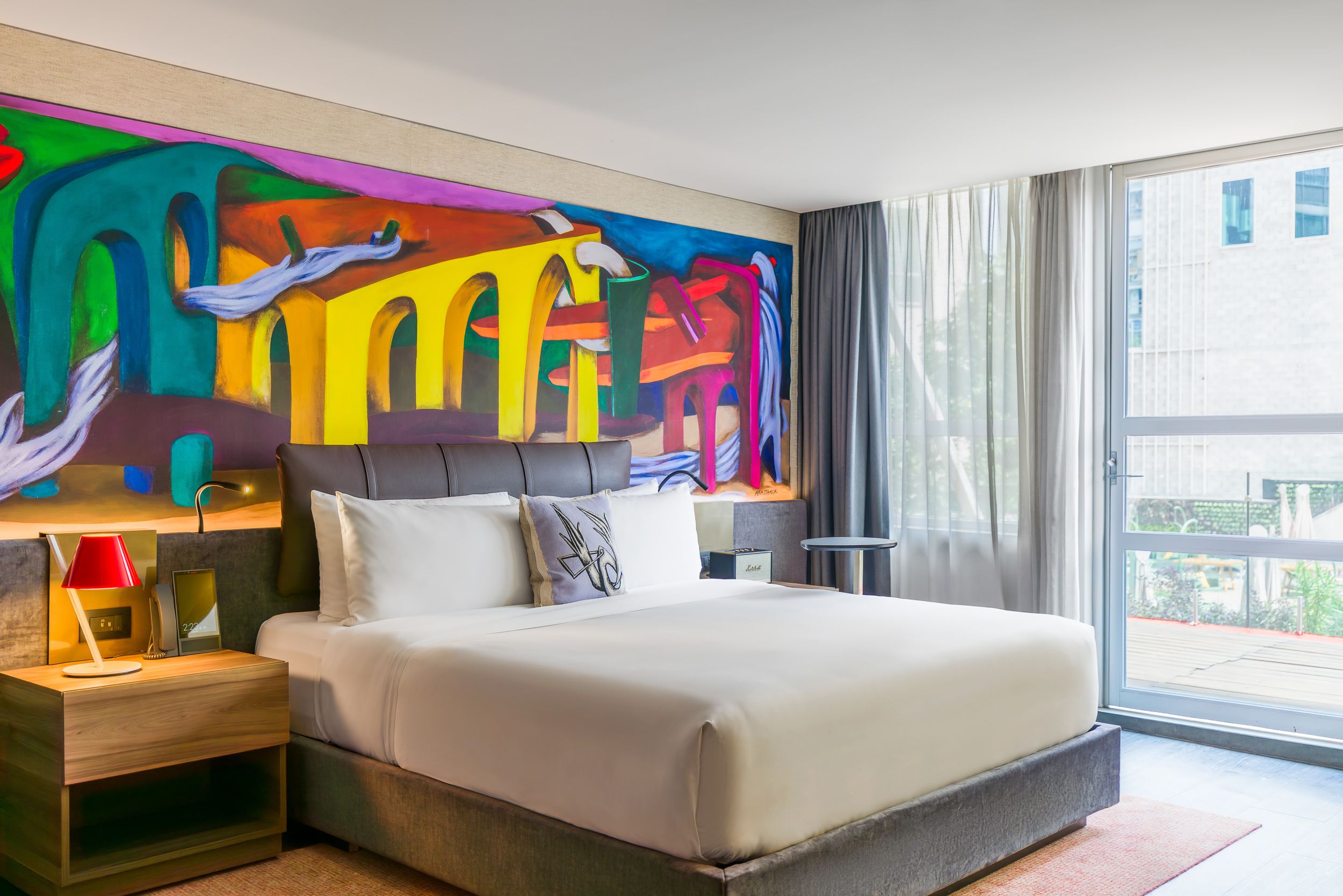 Contemporary hotel bedroom featuring minimalist decor, floor-to-ceiling windows, and bold artistic murals on the walls.