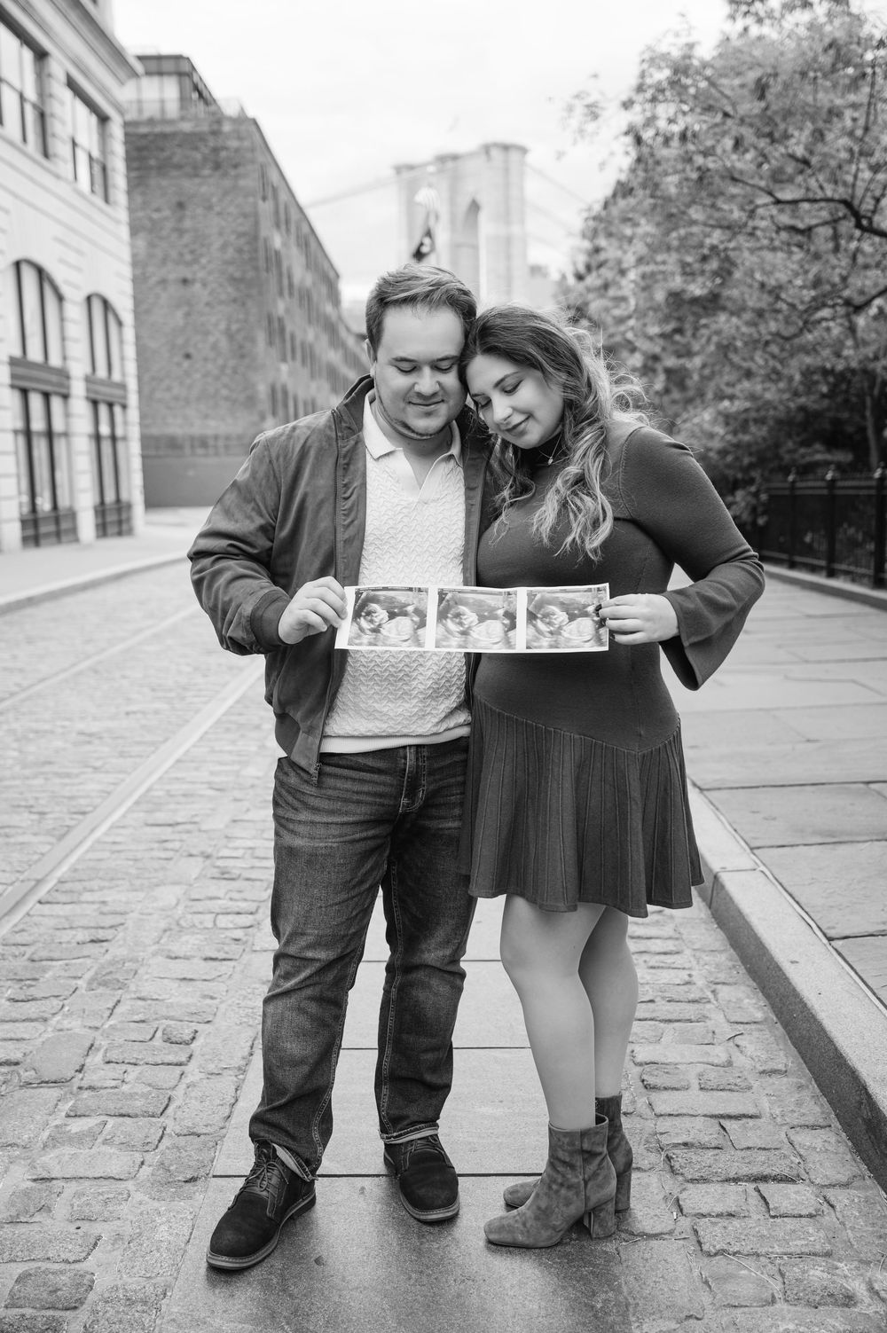 A couple shares an intimate black and white moment on a cobblestone street while holding ultrasound photos.
