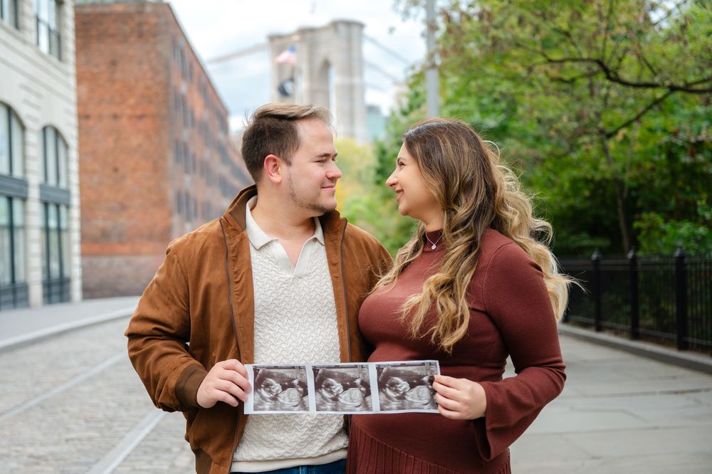 Couple holds an ultrasound photo while standing on cobblestone street with the Brooklyn Bridge in the background during a maternity session.