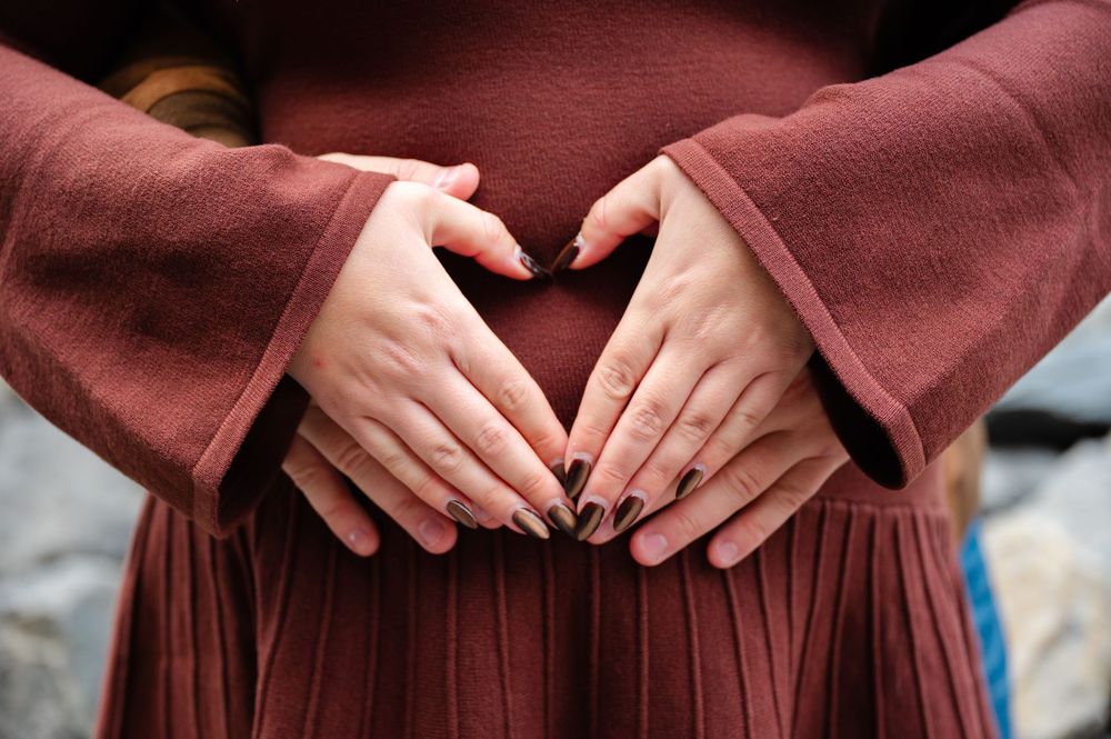 A woman in a burgundy dress holds her pregnant belly while standing in an outdoor setting during sunset.