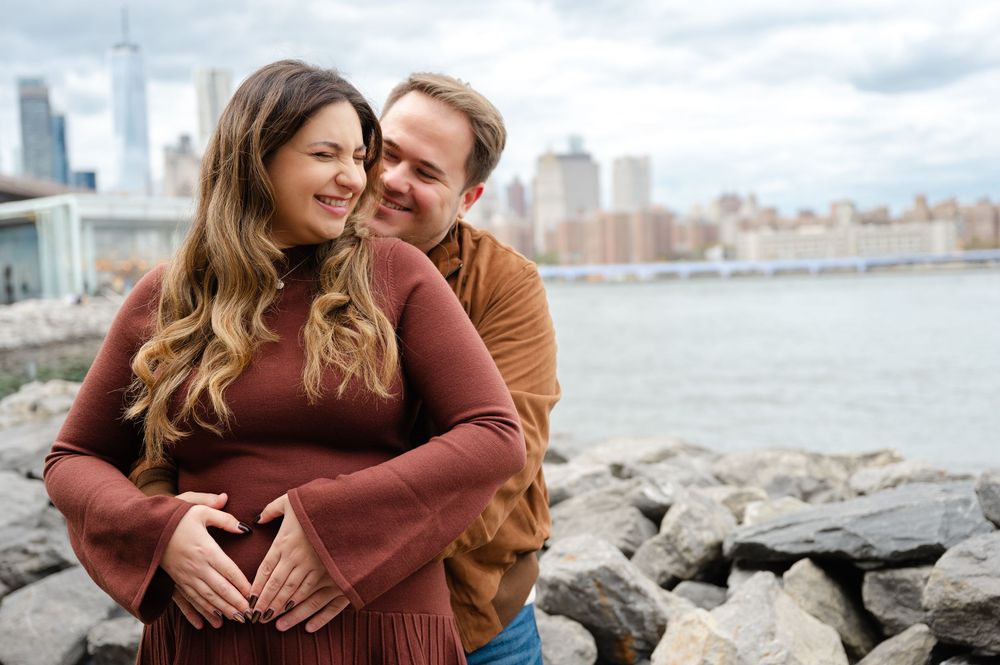 A couple embraces by the rocky shoreline with the Manhattan skyline visible across the water.