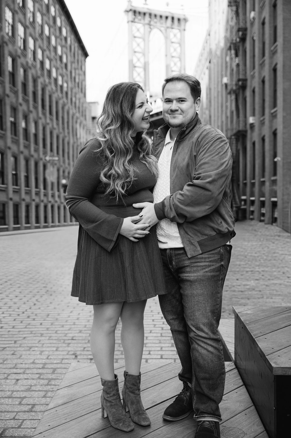 A pregnant woman and her fiancé pose on the cobblestone of Water Street with the Manhattan Bridge visible in the background.