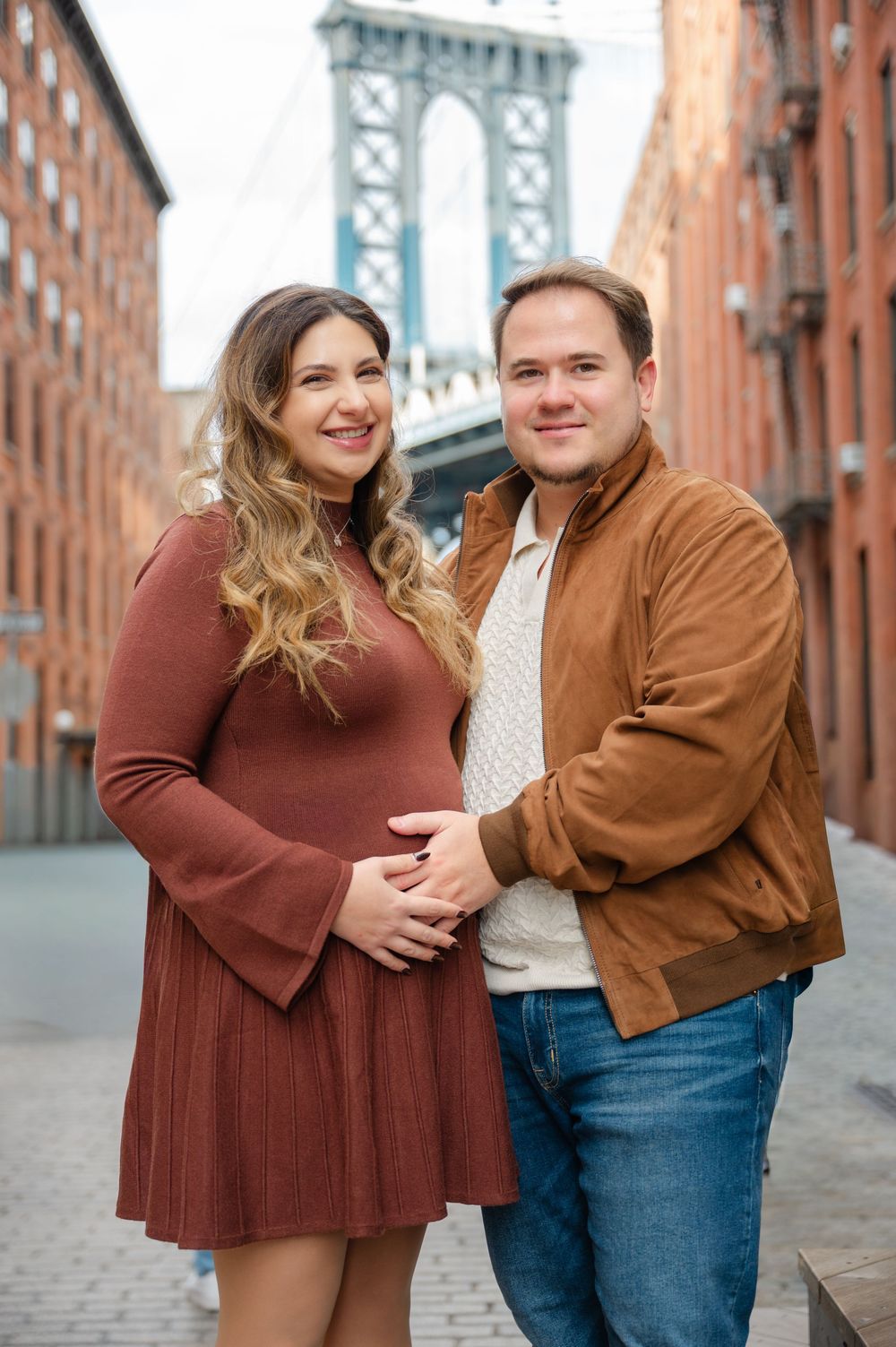 A couple poses playfully together on a cobblestone street with the Manhattan Bridge visible in the background.