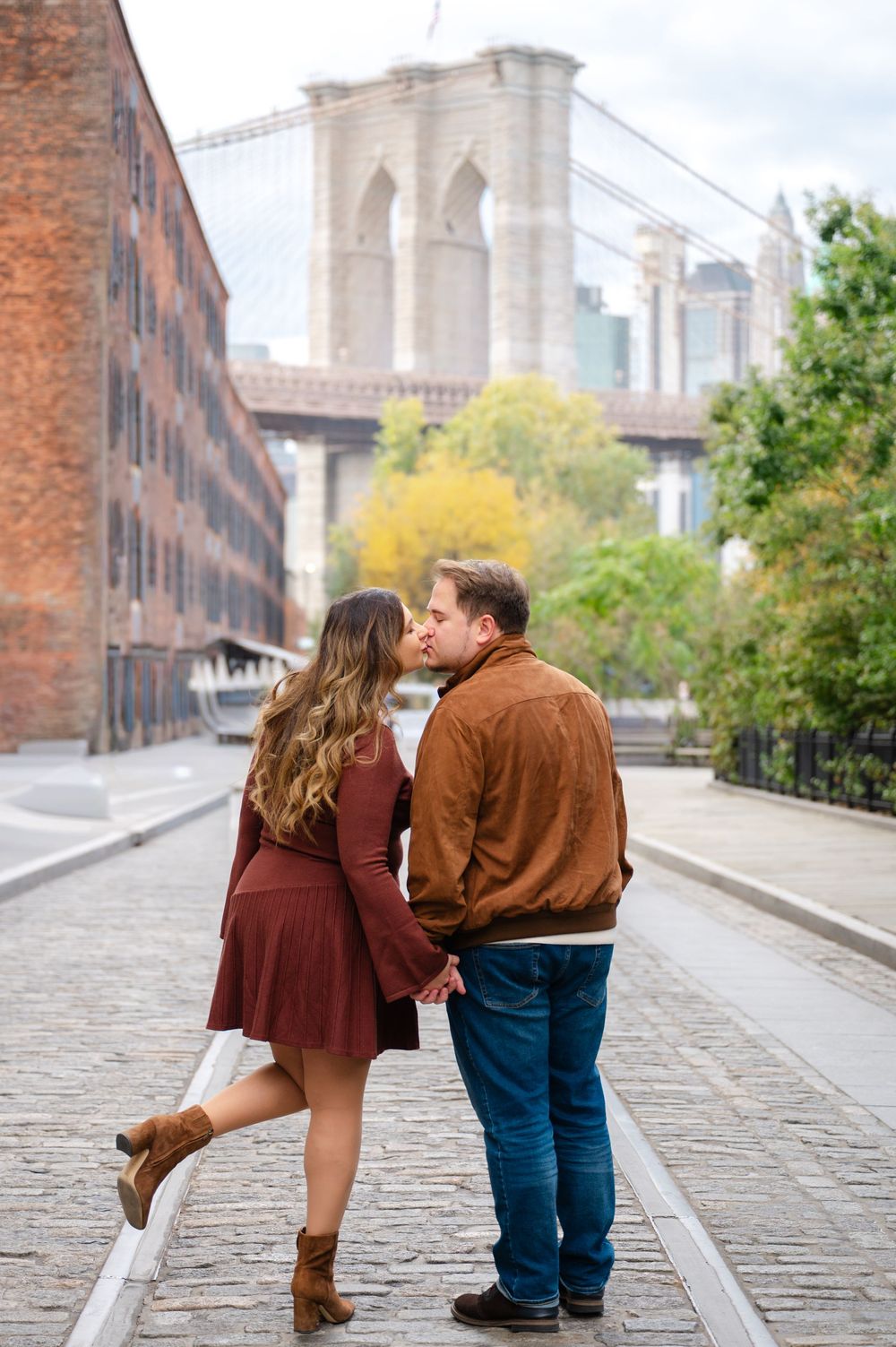 A couple shares a kiss on cobblestone streets with the Brooklyn Bridge and fall foliage in view.