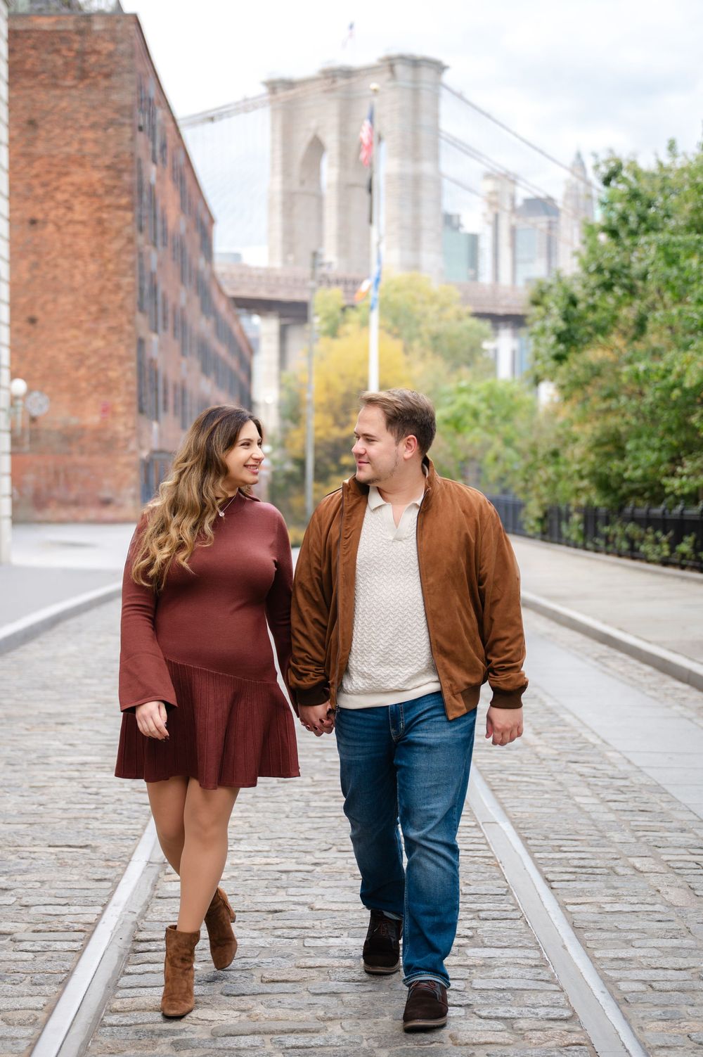 A couple walks hand in hand along a cobblestone street in DUMBO with the Brooklyn Bridge and autumn trees behind them.
