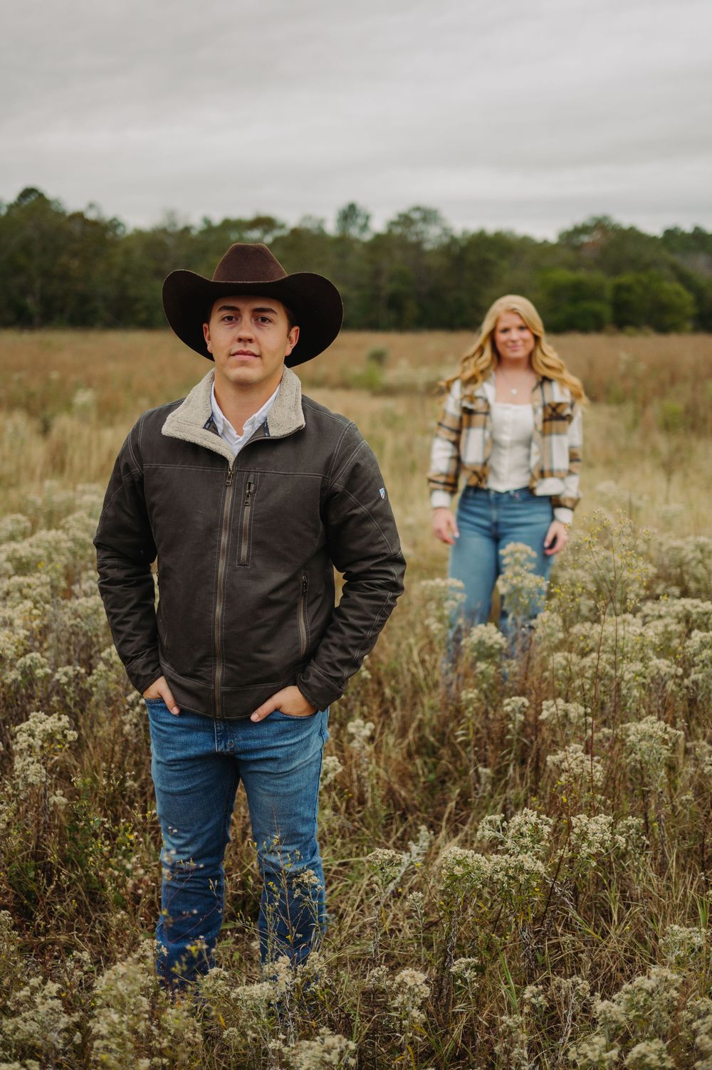 A couple in western attire stands among tall grasses in an open field on an overcast day.
