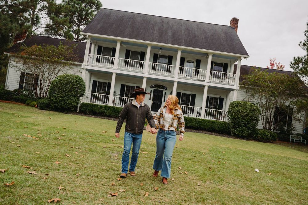 A group walks across a grassy lawn in front of a white colonial-style house with a wraparound porch.