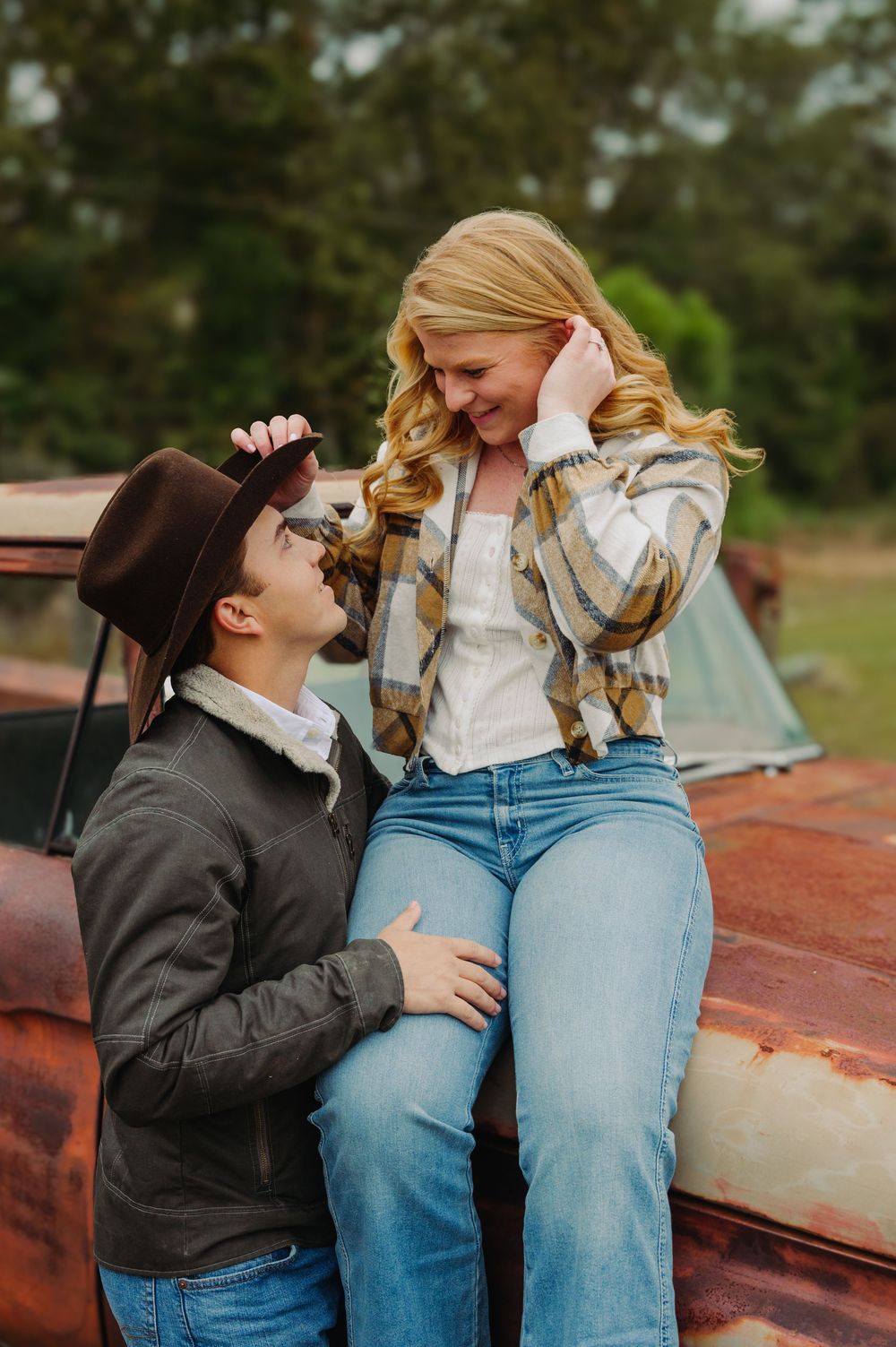 A romantic couple shares a playful moment while sitting on a vintage rusty truck with a camping tent in the background.