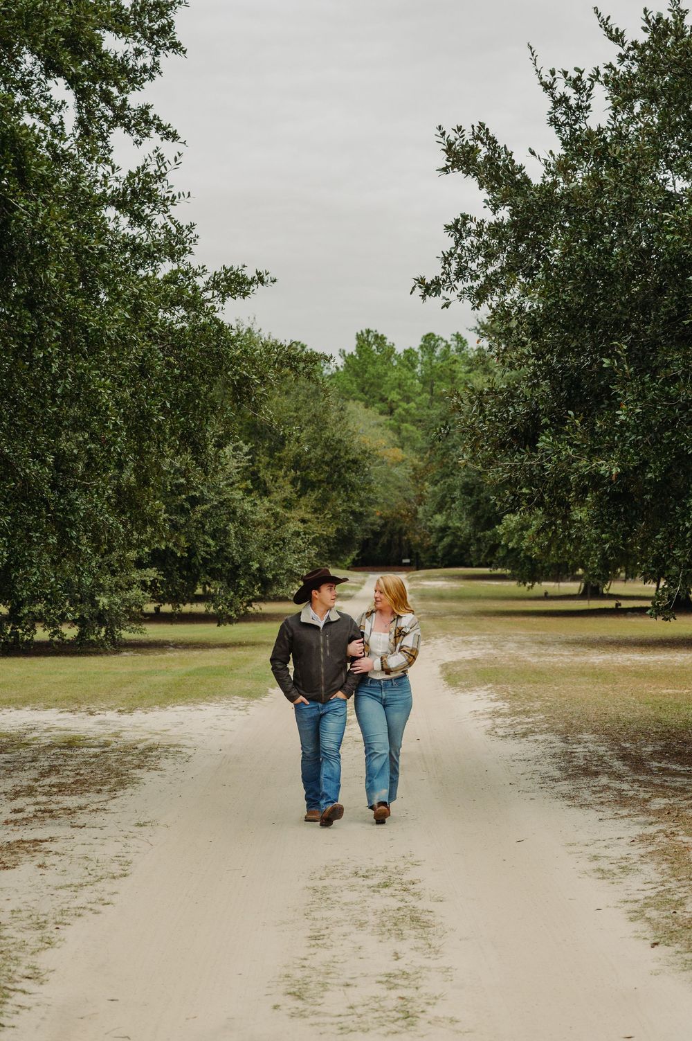 Couple walking hand in hand through a serene orchard with green trees and sandy paths on an overcast day.
