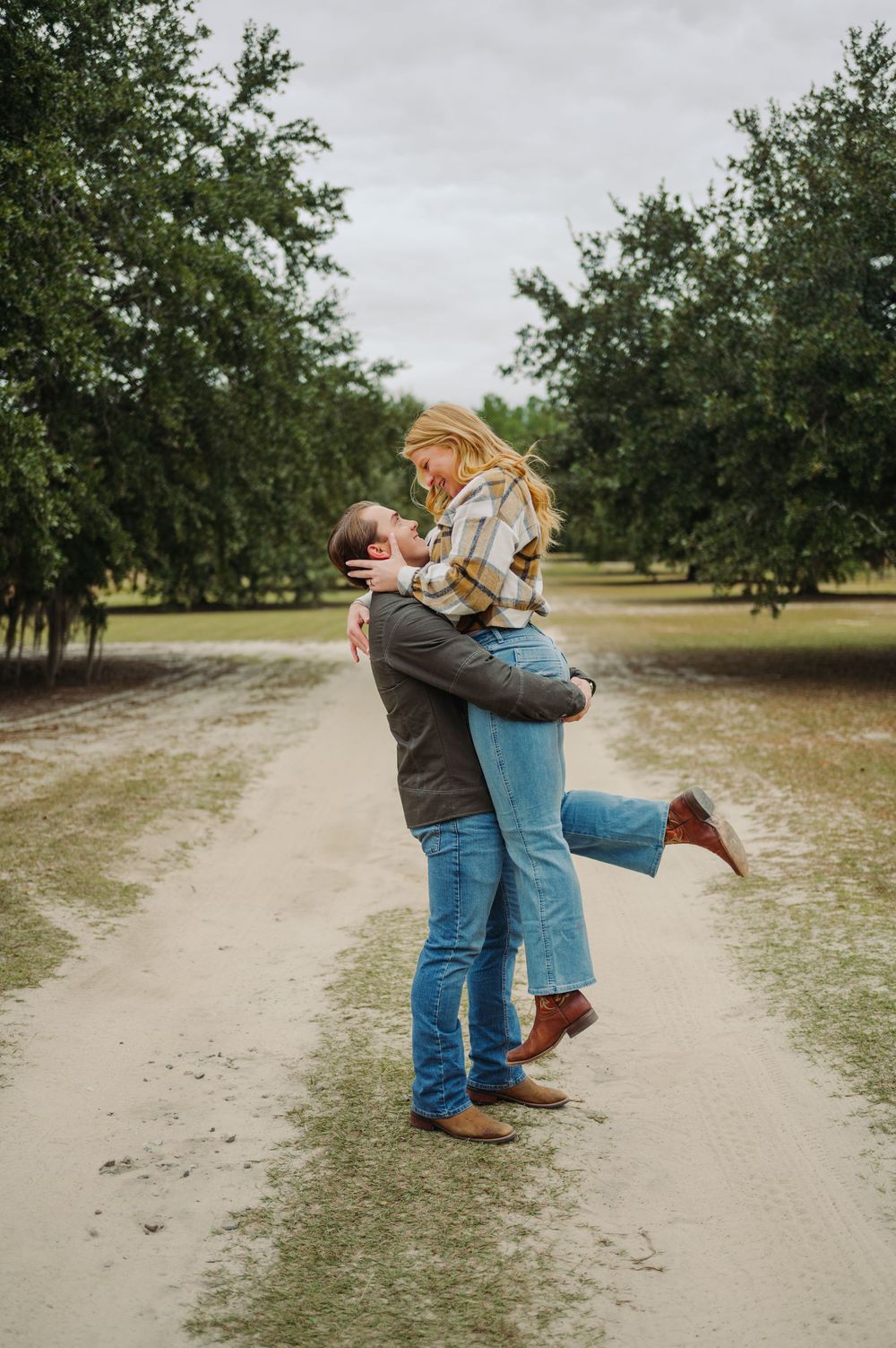 A romantic couple embraces on a tree-lined dirt path, with the woman lifted off her feet in a joyful moment.